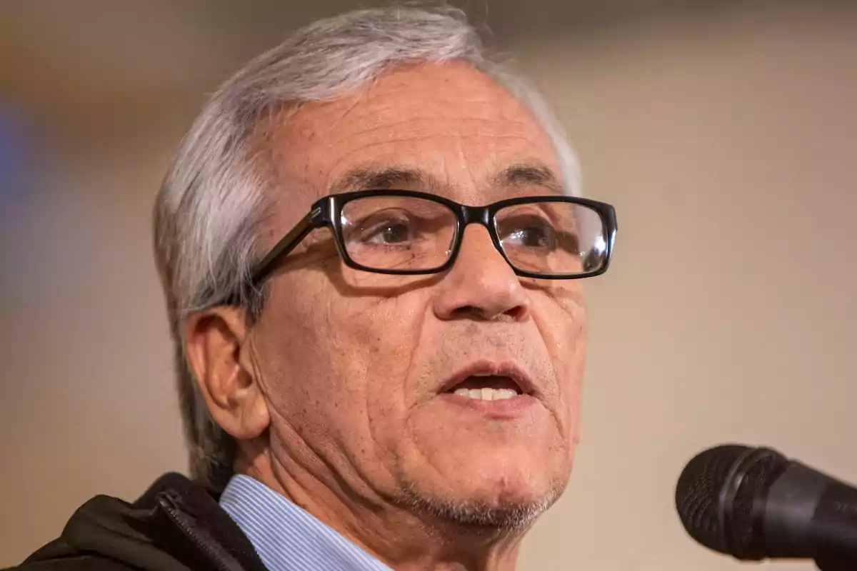 Hombre mayor de cabello canoso y gafas hablando frente a un micrófono