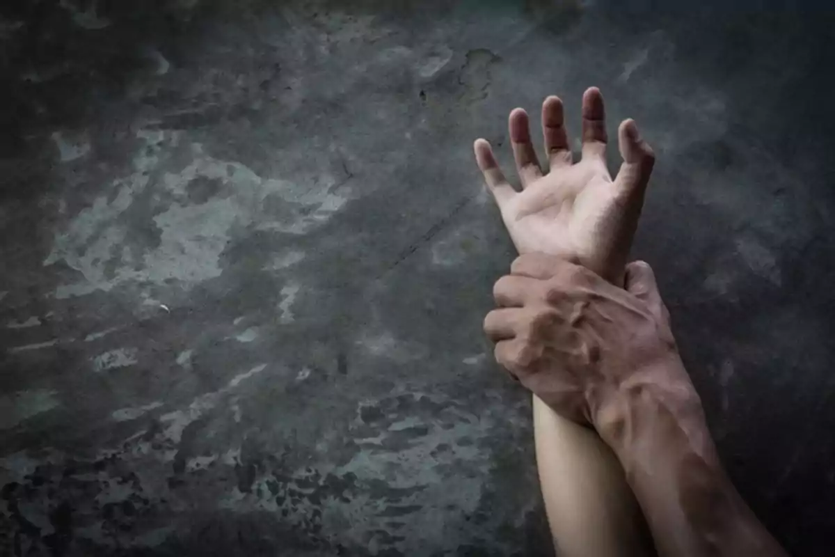 Una mano sujetando con fuerza la muñeca de otra persona sobre una superficie gris