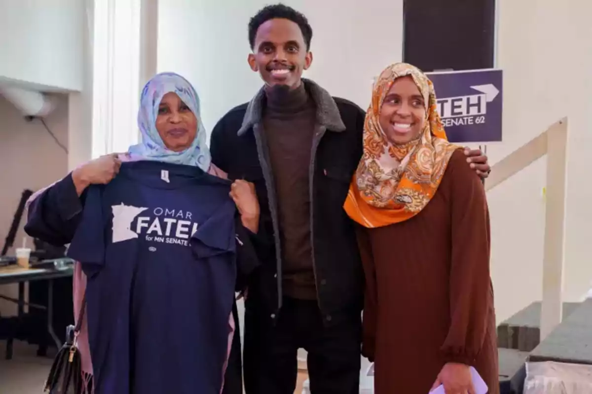 Three people smile at the camera, one of them holding a blue t-jersey with the text Omar Fateh for MN Senate.