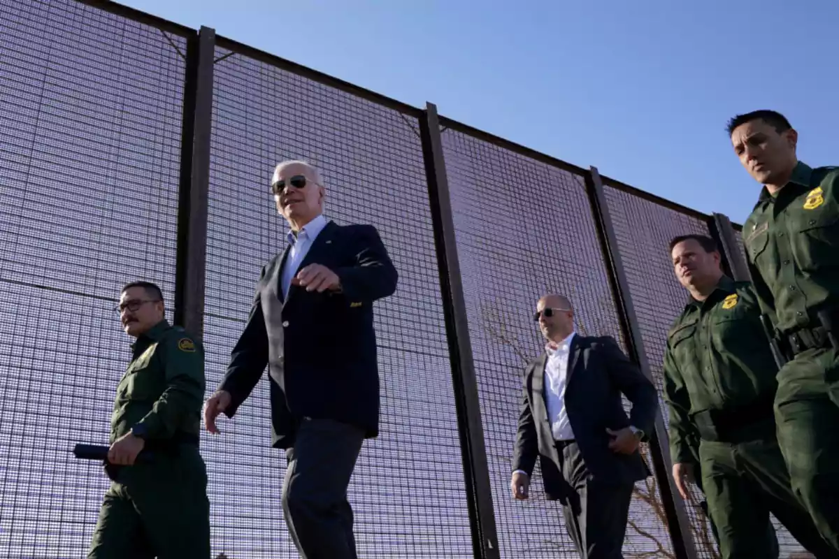 A group of men, some in green uniforms and others in suits, walk next to a tall metal fence under a clear sky. A group of men, some in green uniforms and others in suits, walk next to a tall metal fence under a clear sky.