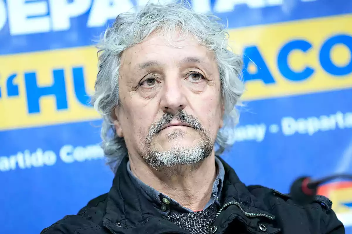 Hombre de cabello canoso y barba mirando al frente con expresión seria frente a un fondo azul y amarillo con letras grandes. Hombre de cabello canoso y barba mirando al frente con expresión seria frente a un fondo azul y amarillo con letras grandes.