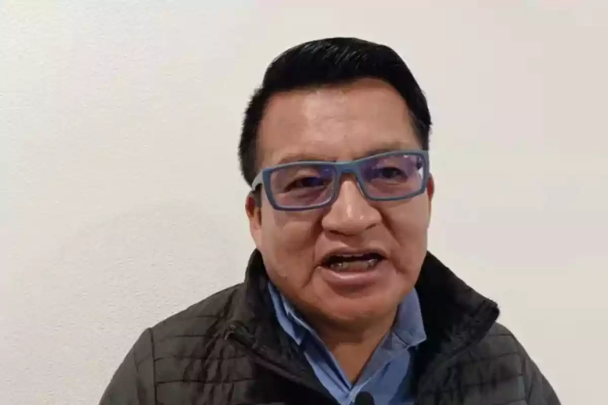 Hombre con gafas azules y chaqueta oscura frente a una pared blanca Hombre con gafas azules y chaqueta oscura frente a una pared blanca