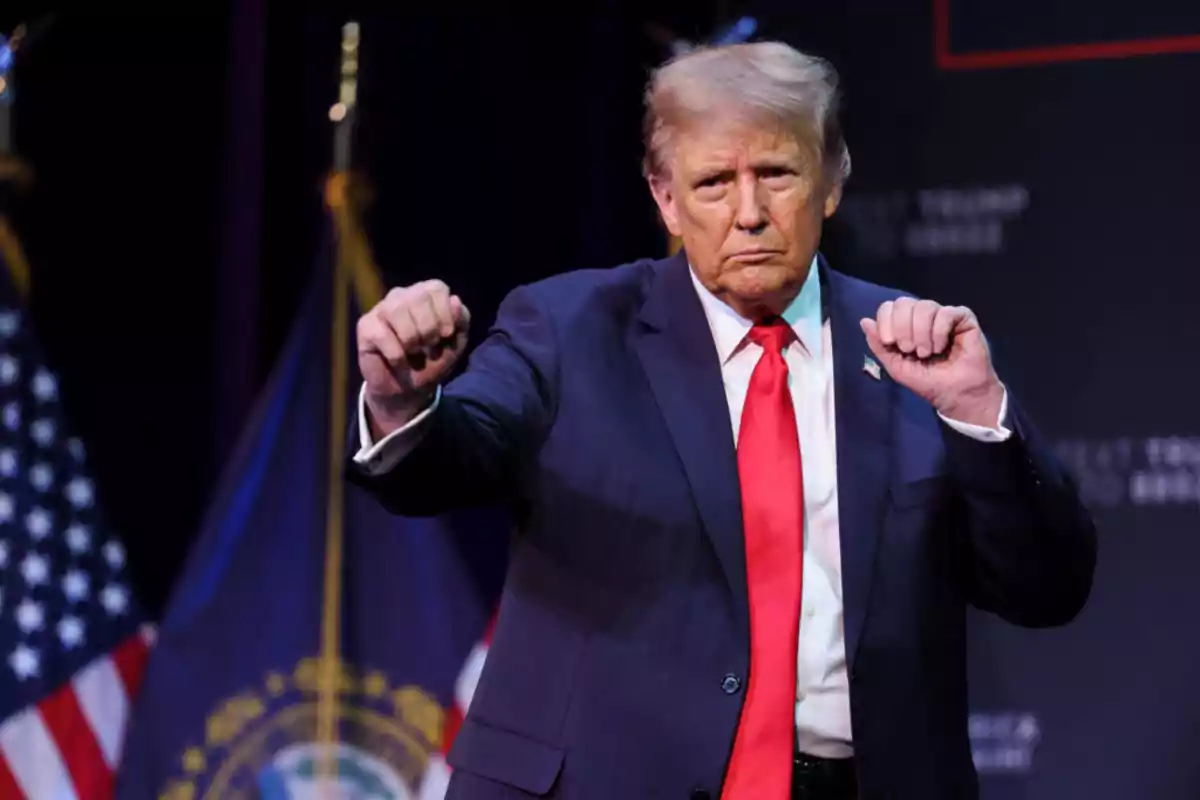 Man in a blue suit and red tie raising both fists on a stage with flags in the background