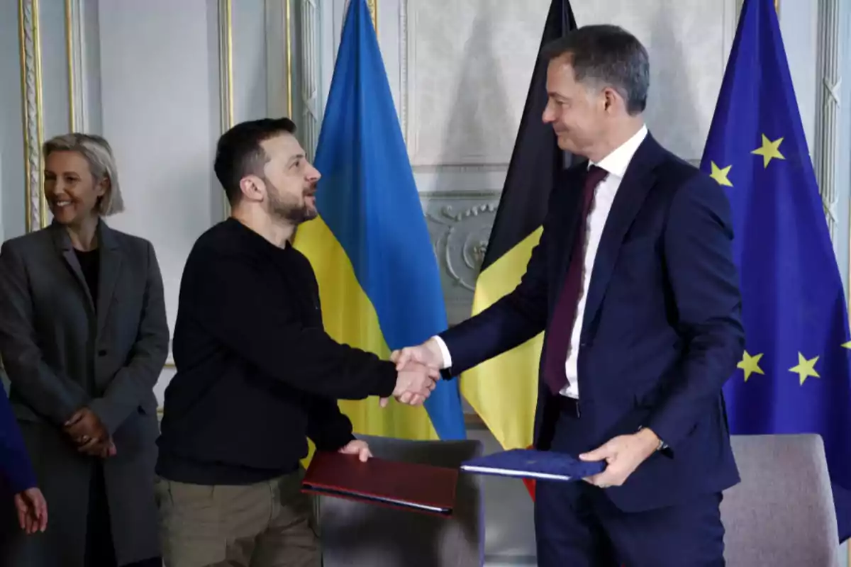 Two men shake hands while holding folders, with the flags of Ukraine, Belgium, and the European Union in the background and a smiling woman watching the scene. Two men shake hands while holding folders, with the flags of Ukraine, Belgium, and the European Union in the background and a smiling woman watching the scene.