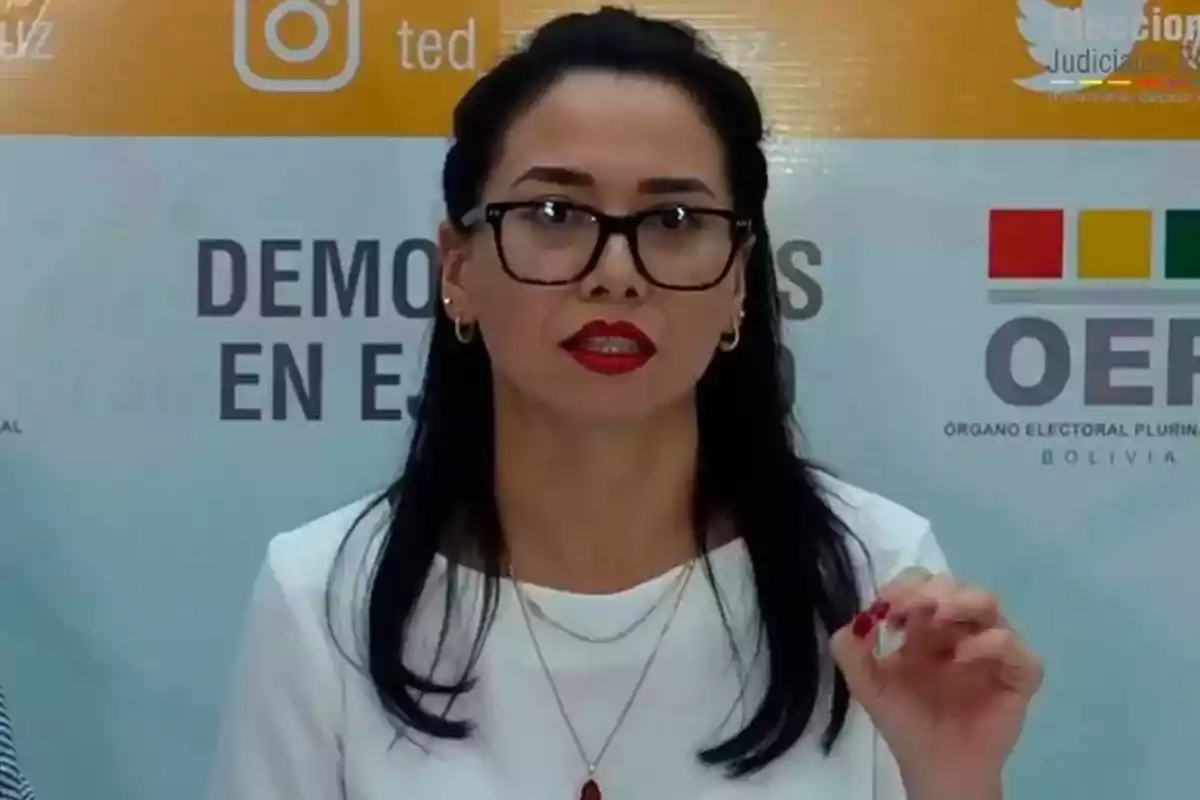 Mujer de cabello oscuro y gafas hablando frente a un fondo con logotipos y texto institucional Mujer de cabello oscuro y gafas hablando frente a un fondo con logotipos y texto institucional