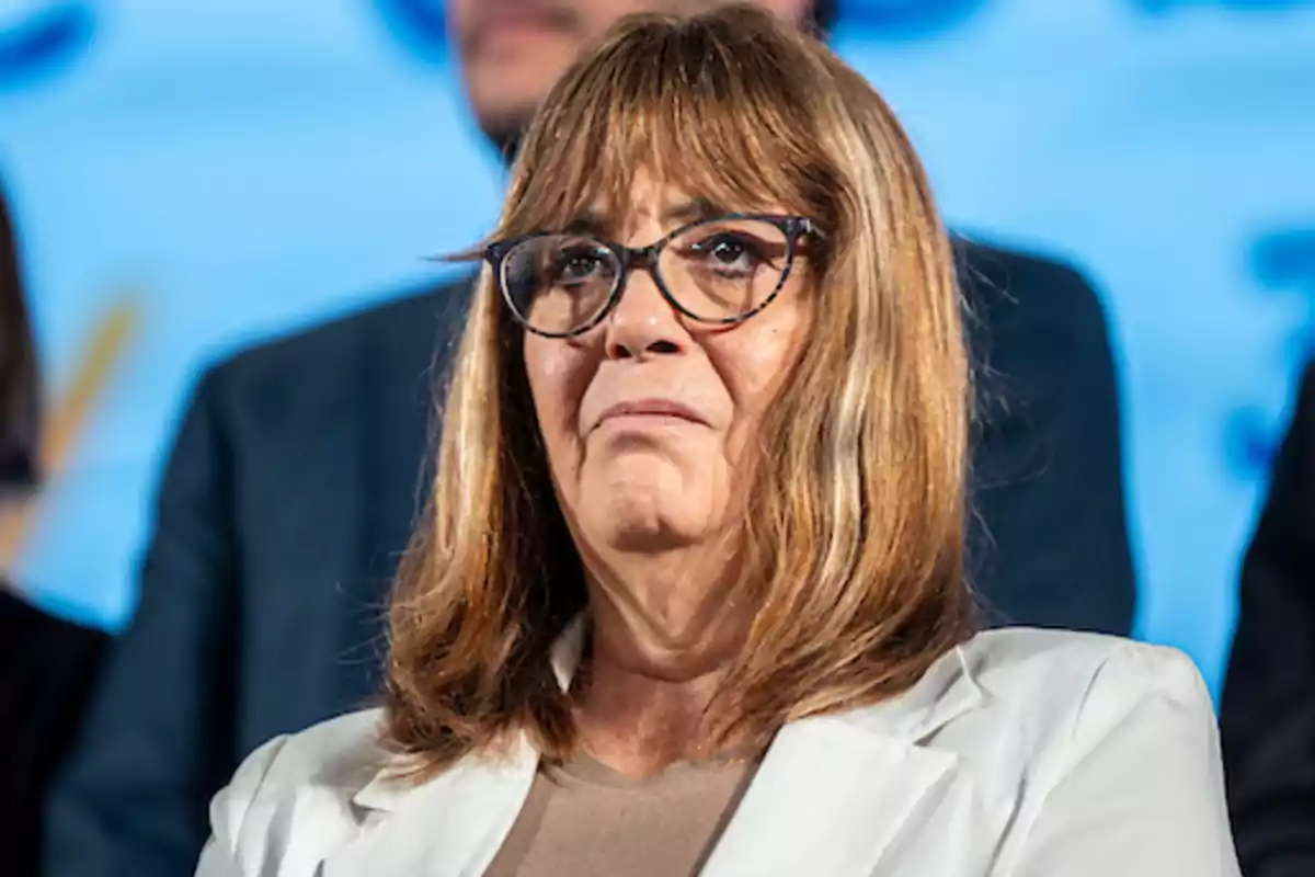 Mujer de cabello castaño y gafas con expresión seria vestida con saco blanco frente a un fondo azul desenfocado