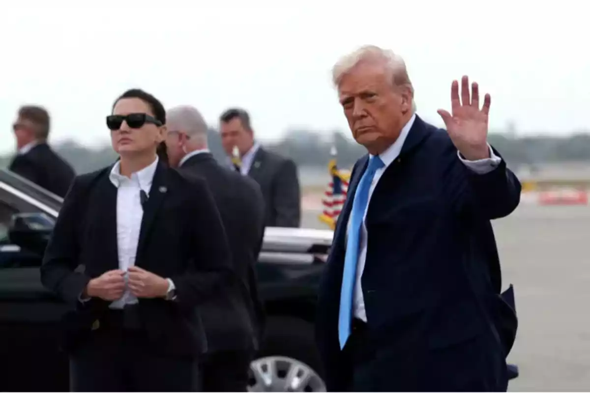 Hombre de cabello rubio con traje azul saludando con la mano mientras una mujer con gafas oscuras y traje oscuro lo acompaña en un entorno al aire libre junto a un vehículo negro