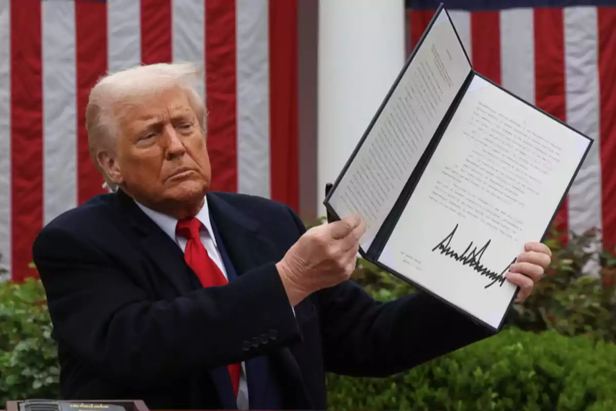 Hombre de cabello rubio y corbata roja sostiene un documento abierto con una firma grande, al fondo hay una bandera de Estados Unidos. Hombre de cabello rubio y corbata roja sostiene un documento abierto con una firma grande, al fondo hay una bandera de Estados Unidos.