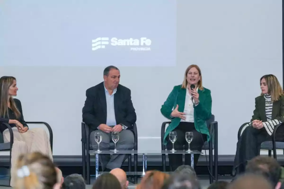Four people seated on a panel in front of an audience with a background displaying the Santa Fe Province logo while a woman speaks holding a microphone. Four people seated on a panel in front of an audience with a background displaying the Santa Fe Province logo while a woman speaks holding a microphone.