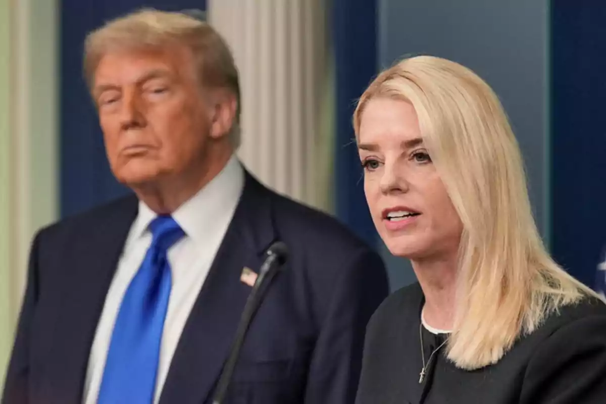 Una mujer rubia habla frente a un micrófono mientras un hombre de cabello rubio y corbata azul la observa en segundo plano