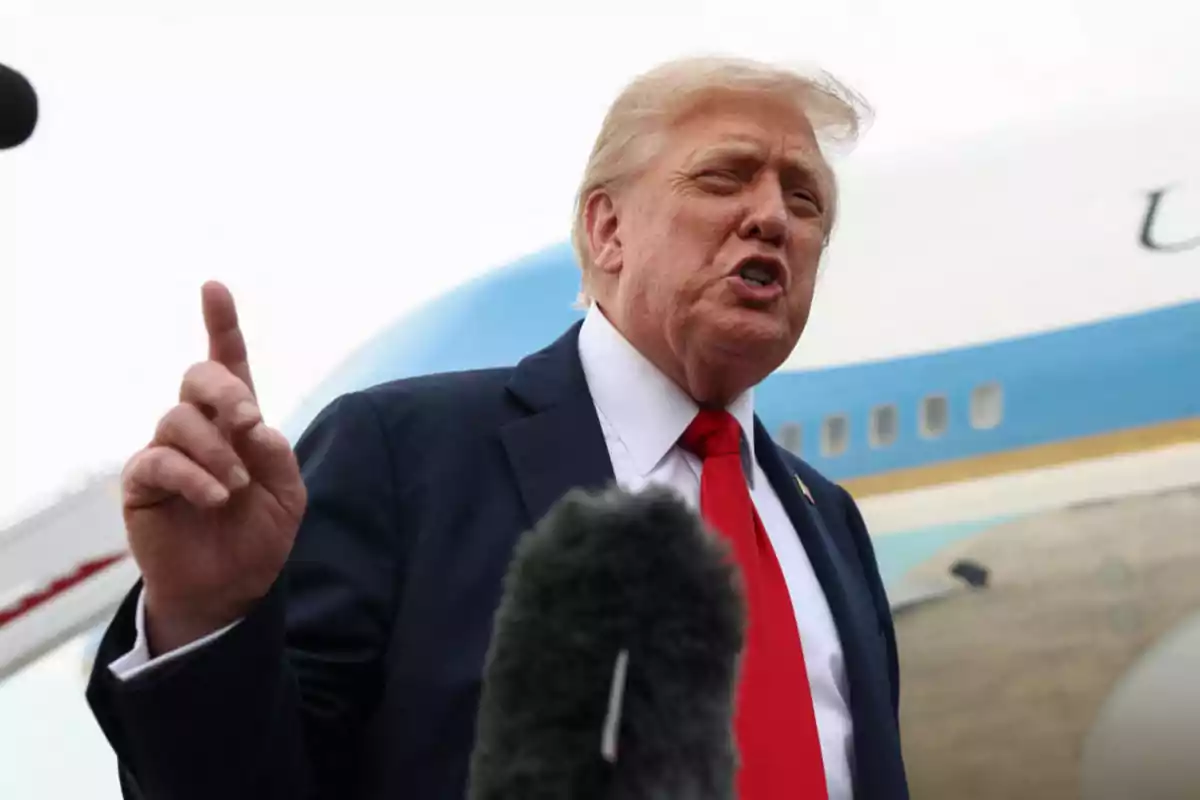 Blond-haired man in a dark suit and red tie speaking in front of a microphone with an airplane in the background Blond-haired man in a dark suit and red tie speaking in front of a microphone with an airplane in the background