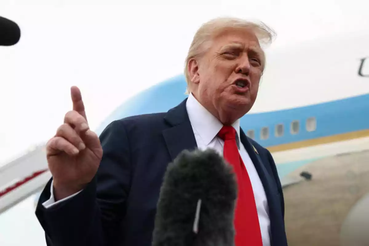 Blond-haired man in a dark suit and red tie speaking in front of a microphone with an airplane in the background