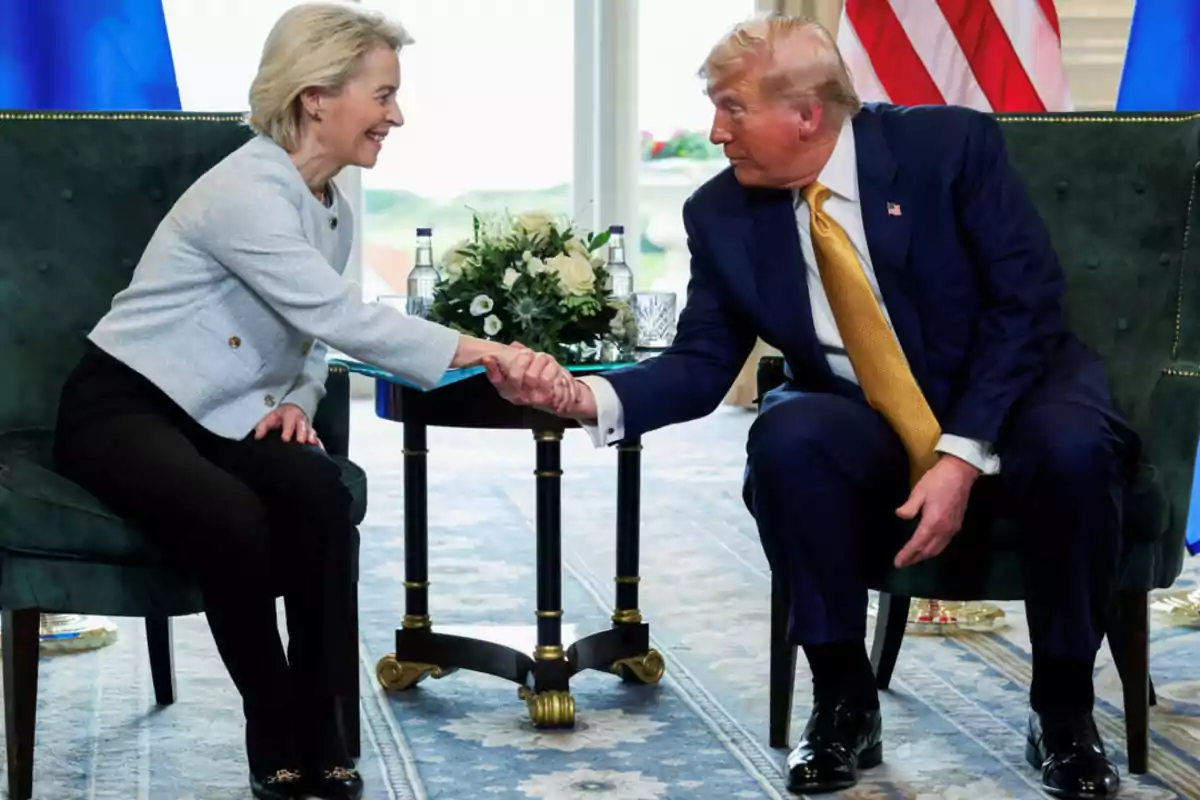 Dos personas elegantemente vestidas se dan la mano mientras están sentadas frente a frente en una sala con banderas y flores de fondo Dos personas elegantemente vestidas se dan la mano mientras están sentadas frente a frente en una sala con banderas y flores de fondo