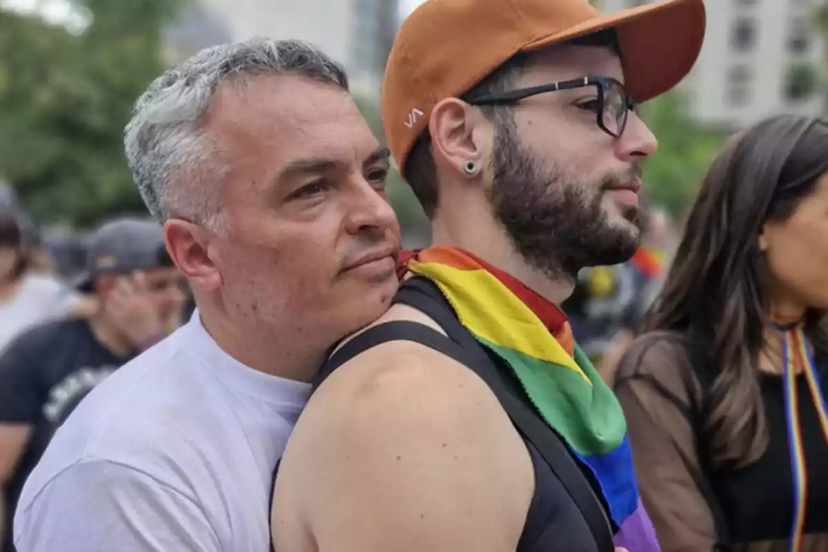 Dos hombres abrazados en un evento al aire libre uno de ellos lleva una bandera arcoíris alrededor del cuello y una gorra naranja