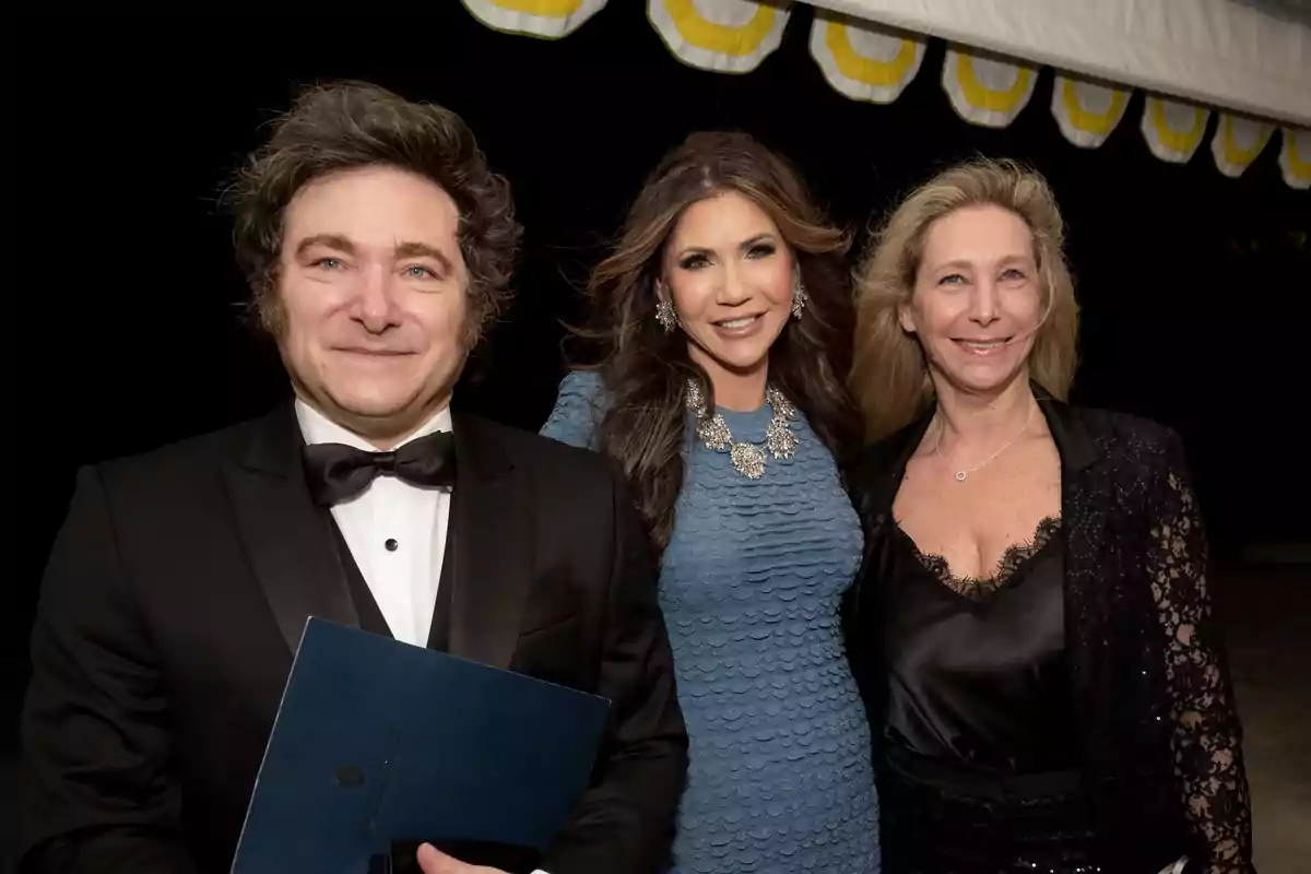 Tres personas posan sonrientes en un evento elegante, el hombre lleva esmoquin y las dos mujeres visten ropa formal Tres personas posan sonrientes en un evento elegante, el hombre lleva esmoquin y las dos mujeres visten ropa formal