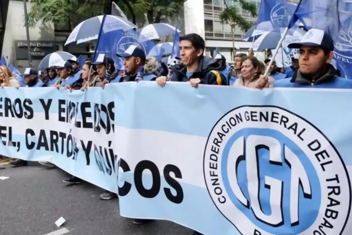 Personas marchando en la calle con una gran bandera celeste y blanca de la Confederación General del Trabajo CGT y paraguas azules