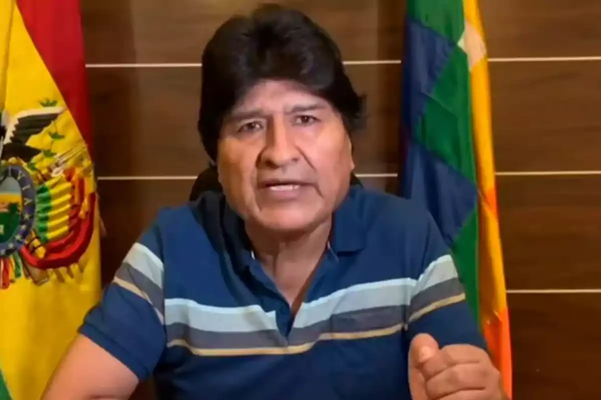 Hombre de cabello oscuro con camiseta azul hablando frente a dos banderas y un fondo de madera Hombre de cabello oscuro con camiseta azul hablando frente a dos banderas y un fondo de madera