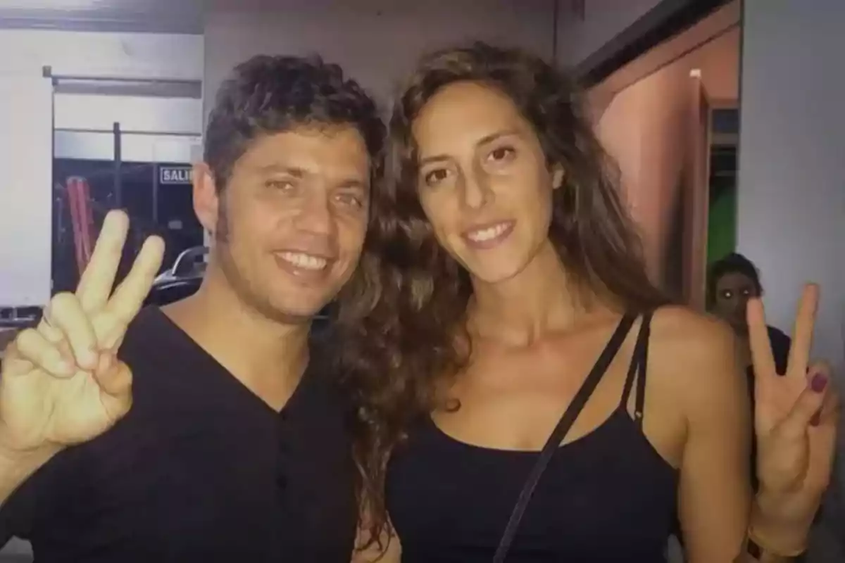 A couple smiles and makes the peace sign with their fingers while posing together indoors.