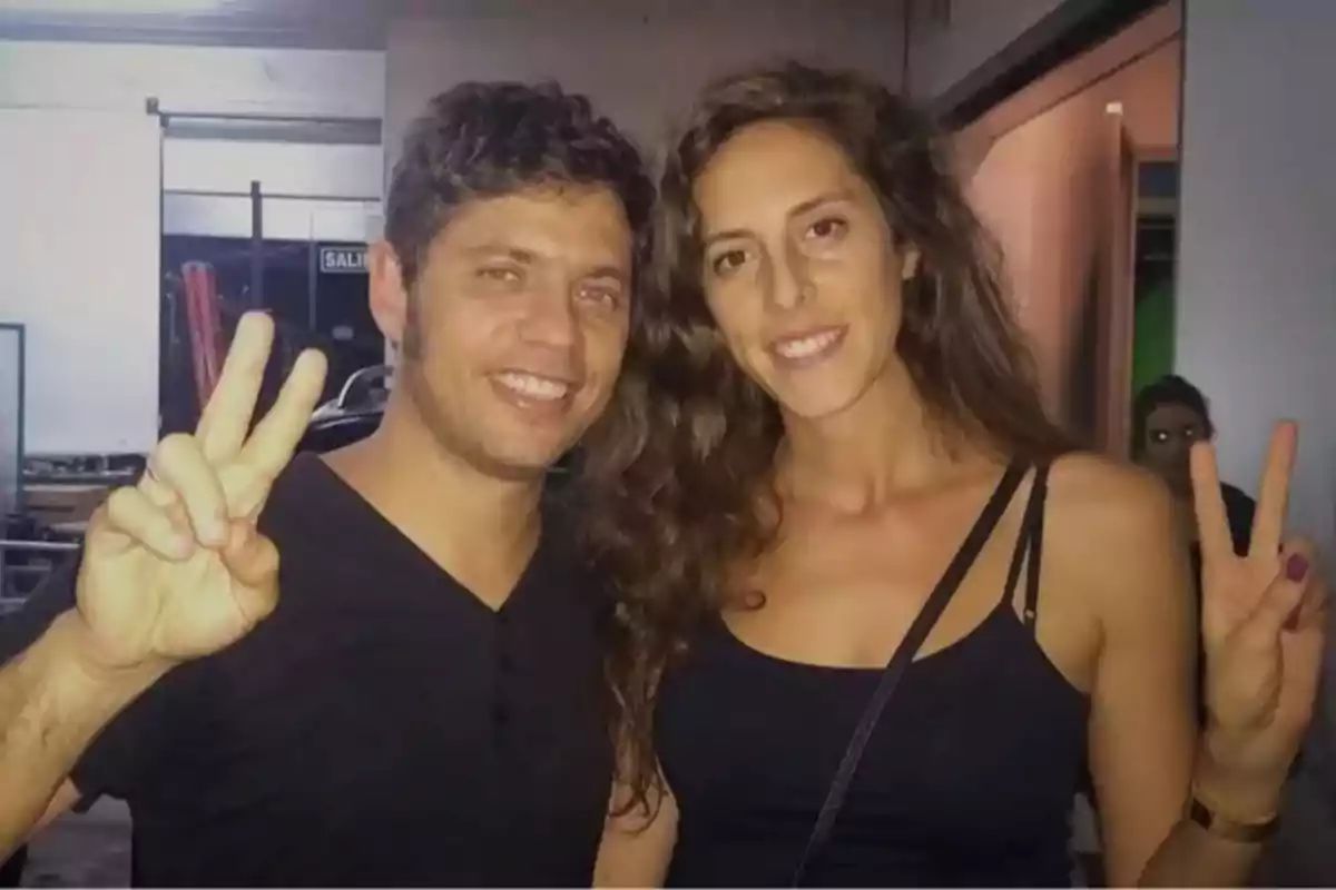 Dos personas sonríen y posan haciendo el gesto de la paz con los dedos en un ambiente interior