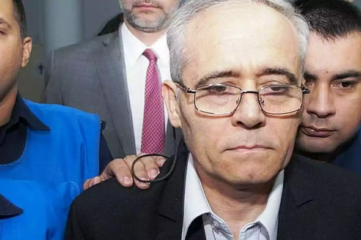 Hombre de cabello canoso y gafas escoltado por varias personas en un ambiente formal Hombre de cabello canoso y gafas escoltado por varias personas en un ambiente formal