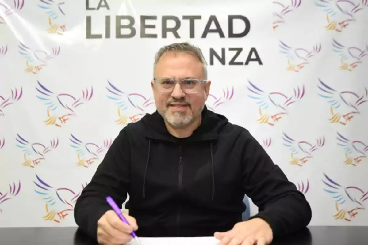 Man with glasses and a beard sitting at a table writing with a purple pen, in the background a poster with the word Man with glasses and a beard sitting at a table writing with a purple pen, in the background a poster with the word