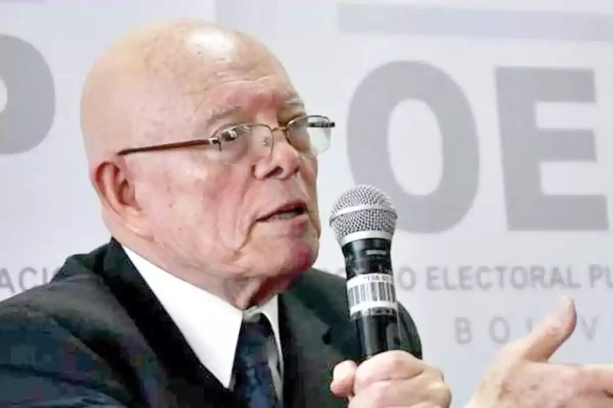 Hombre mayor con gafas y traje hablando en un micrófono durante un evento oficial
