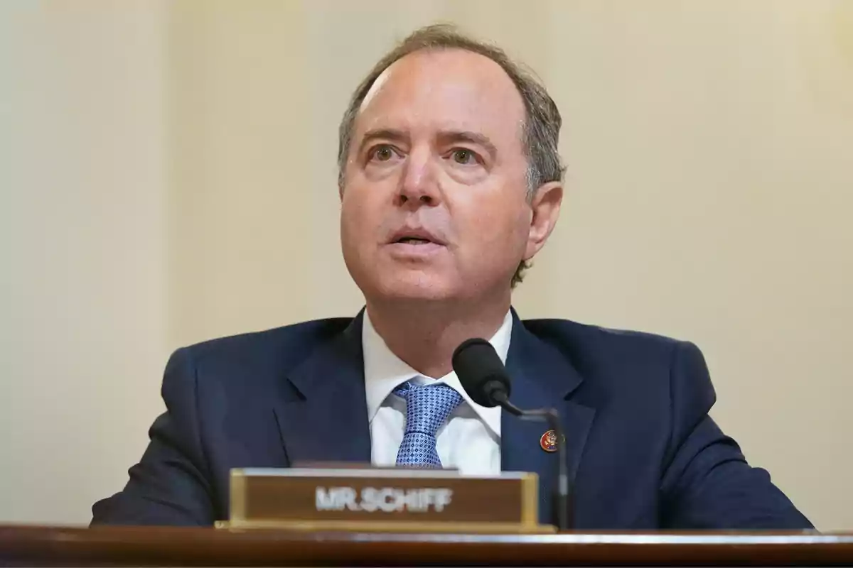 Hombre de traje azul sentado frente a un micrófono con una placa que dice MR SCHIFF Hombre de traje azul sentado frente a un micrófono con una placa que dice MR SCHIFF