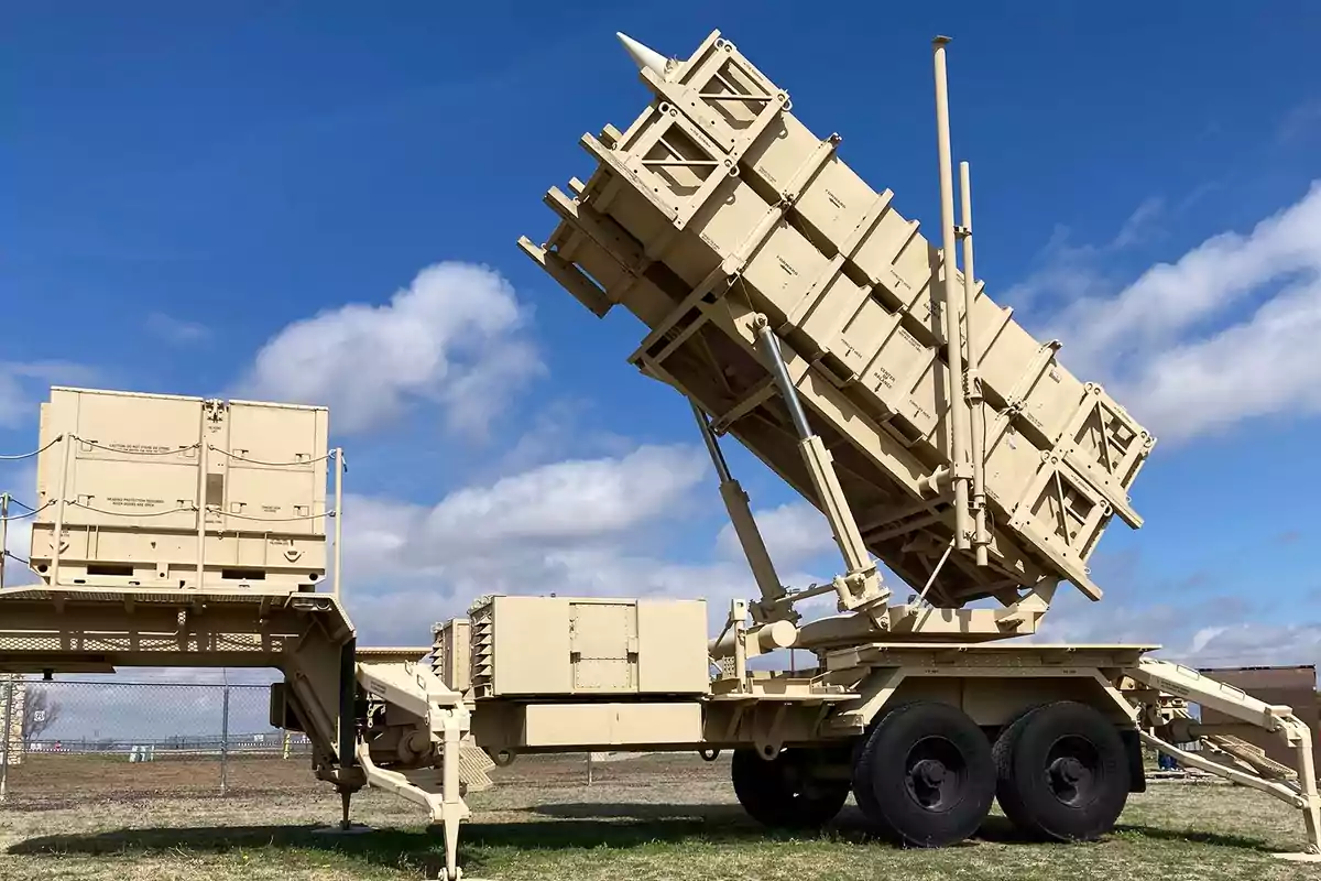 Lanzador de misiles militar color beige en posición de disparo sobre un remolque en un campo bajo un cielo azul con nubes