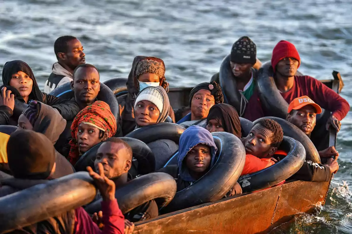 A group of migrants travels on a boat across the sea; many of them are using tires as flotation devices and look toward the camera with serious expressions. A group of migrants travels on a boat across the sea; many of them are using tires as flotation devices and look toward the camera with serious expressions.