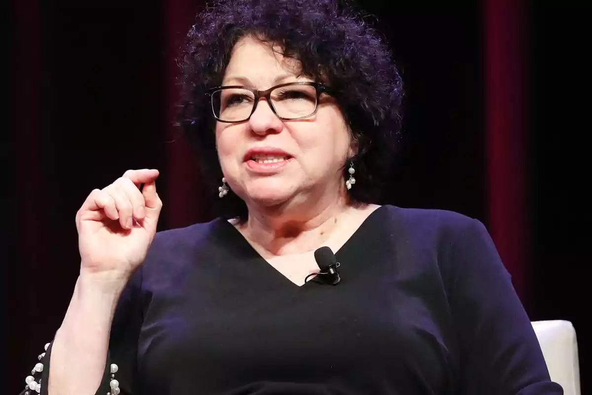 Mujer de cabello rizado y oscuro con gafas y aretes, vestida de negro, hablando y gesticulando mientras está sentada en un escenario Mujer de cabello rizado y oscuro con gafas y aretes, vestida de negro, hablando y gesticulando mientras está sentada en un escenario