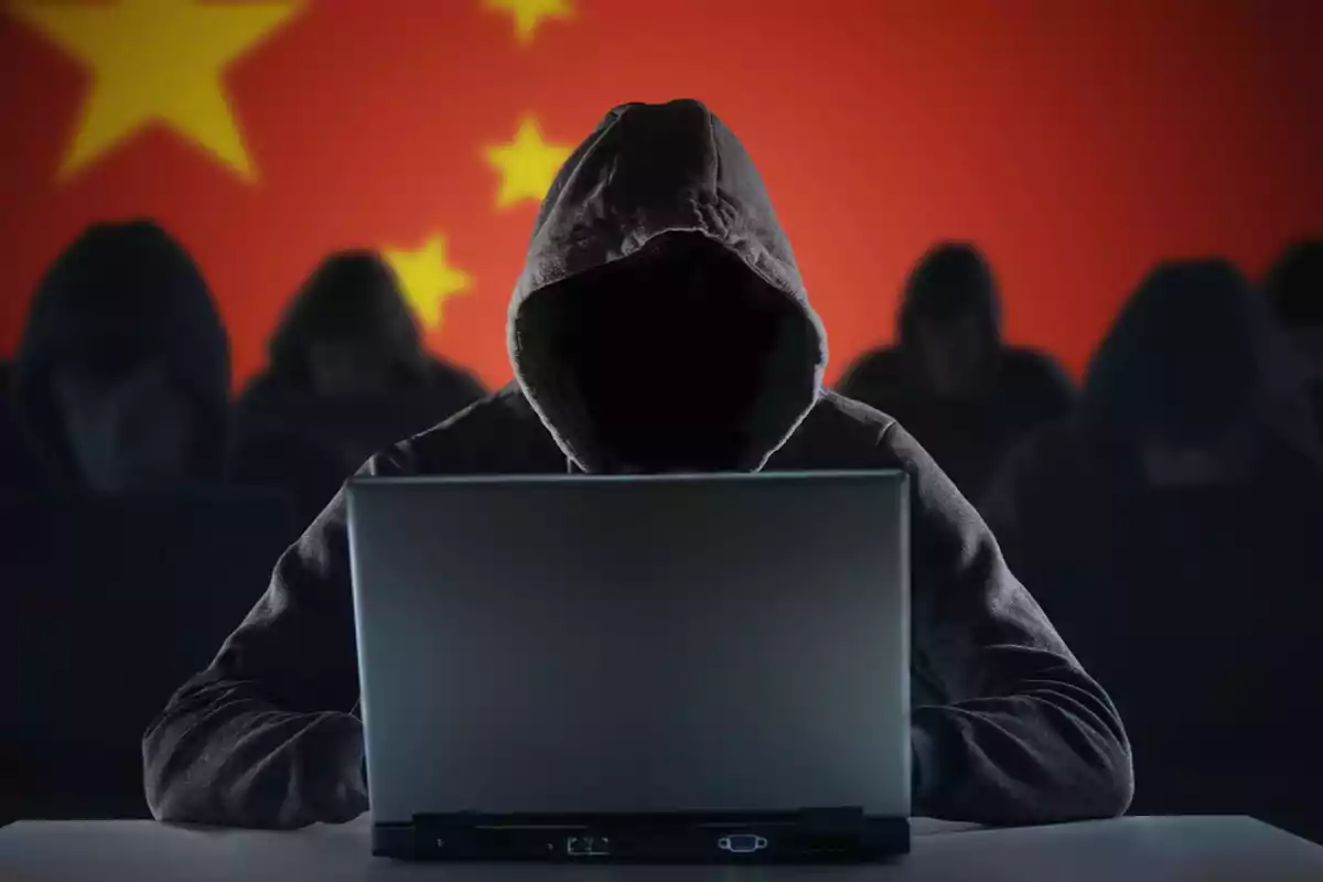 Hooded person using a laptop in front of a Chinese flag with other similar figures in the background