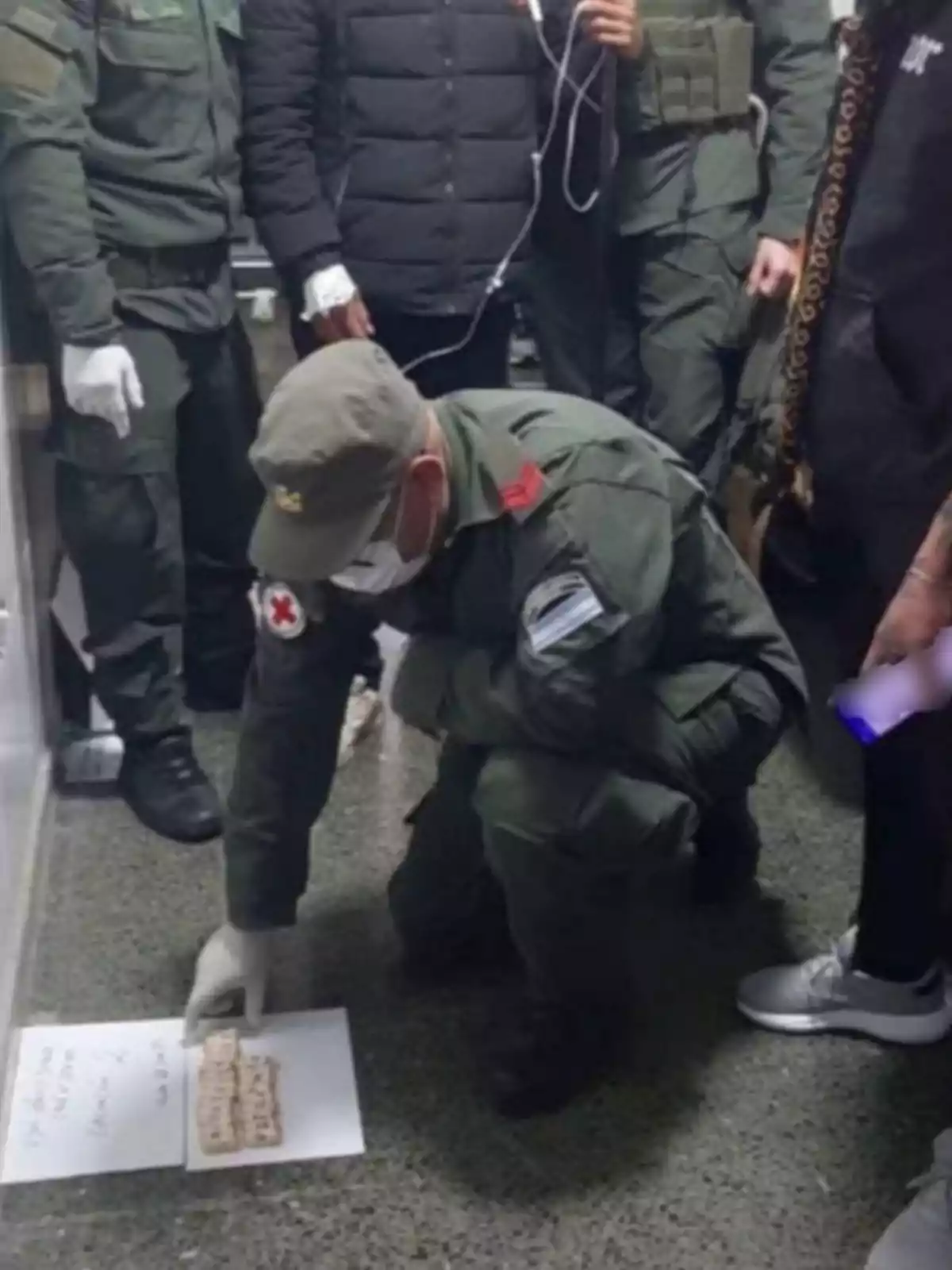 Un grupo de personas con uniforme militar observa mientras una persona se agacha y señala unas galletas sobre una hoja de papel en el suelo Un grupo de personas con uniforme militar observa mientras una persona se agacha y señala unas galletas sobre una hoja de papel en el suelo