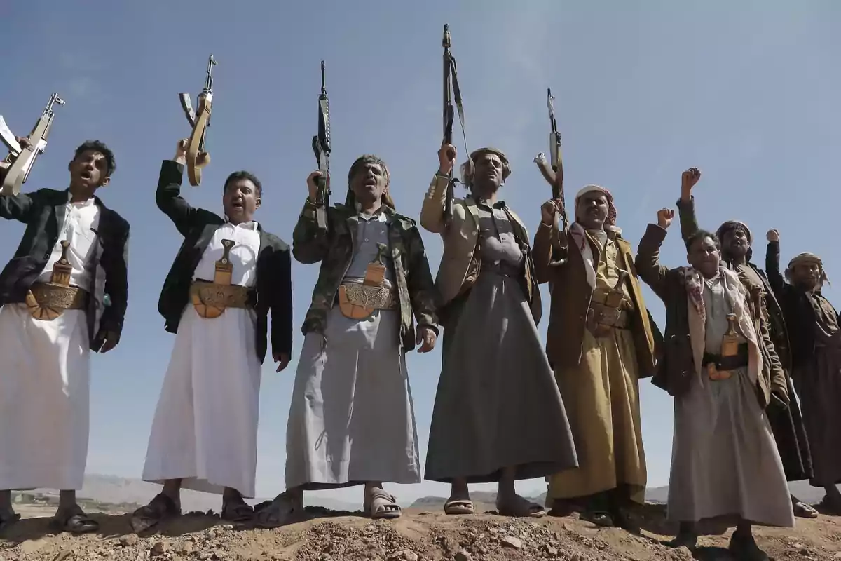 Group of men dressed in traditional Yemeni clothing, some carrying weapons and raising their arms, standing on arid ground under a clear sky. Group of men dressed in traditional Yemeni clothing, some carrying weapons and raising their arms, standing on arid ground under a clear sky.