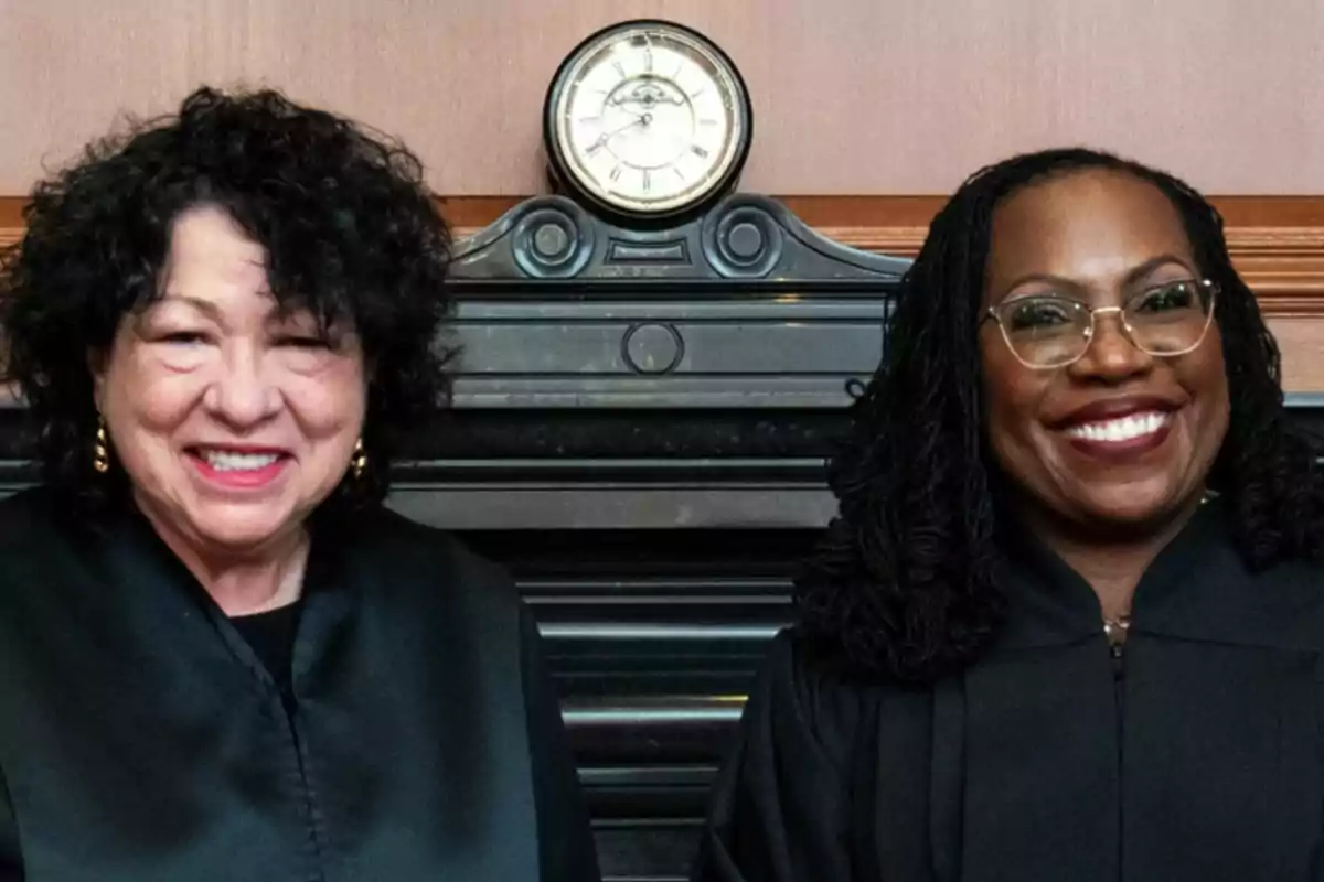 Two women in black robes smile in front of a fireplace with an antique clock on top. Two women in black robes smile in front of a fireplace with an antique clock on top.