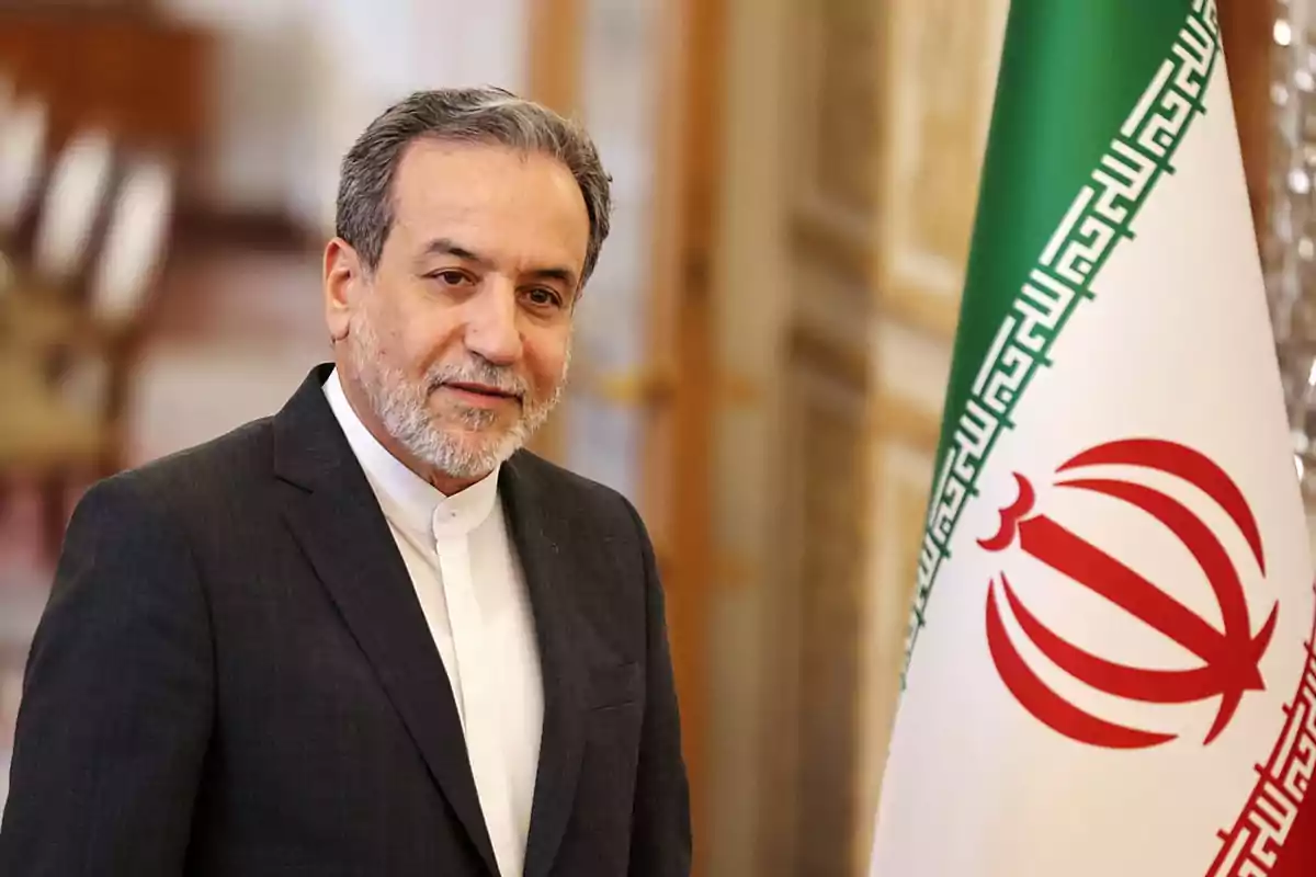 Hombre de cabello canoso y barba con traje oscuro junto a una bandera de Irán en un entorno formal Hombre de cabello canoso y barba con traje oscuro junto a una bandera de Irán en un entorno formal