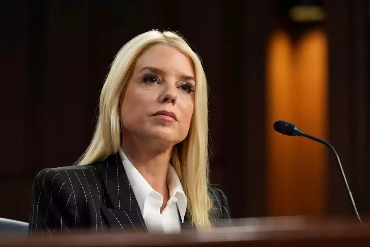 Blonde woman with a dark blazer and white jersey sitting in front of a microphone in a formal setting Blonde woman with a dark blazer and white jersey sitting in front of a microphone in a formal setting