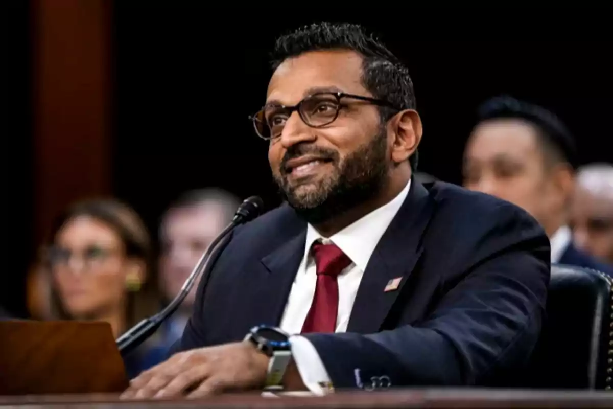 Man with a beard and glasses smiling while speaking into a microphone during a formal hearing
