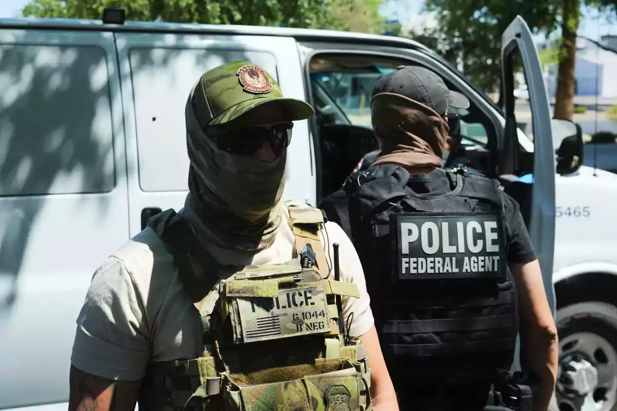 Two federal agents wearing bulletproof vests and with their faces covered stand next to a white van during an outdoor police operation.