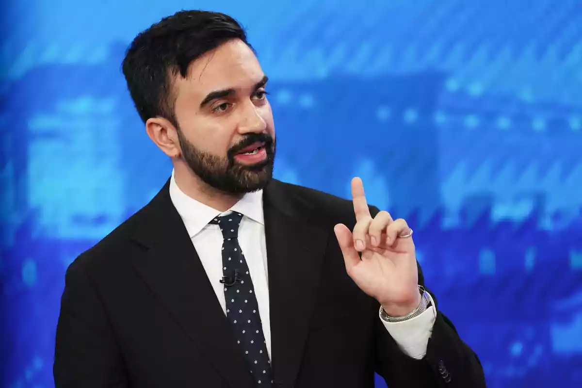 Man with a beard and dark suit speaking and raising a finger in front of a blue background