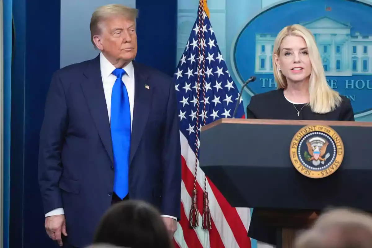 Un hombre con traje azul y corbata azul de pie junto a una mujer rubia hablando en un podio con el escudo presidencial de Estados Unidos y una bandera estadounidense de fondo Un hombre con traje azul y corbata azul de pie junto a una mujer rubia hablando en un podio con el escudo presidencial de Estados Unidos y una bandera estadounidense de fondo