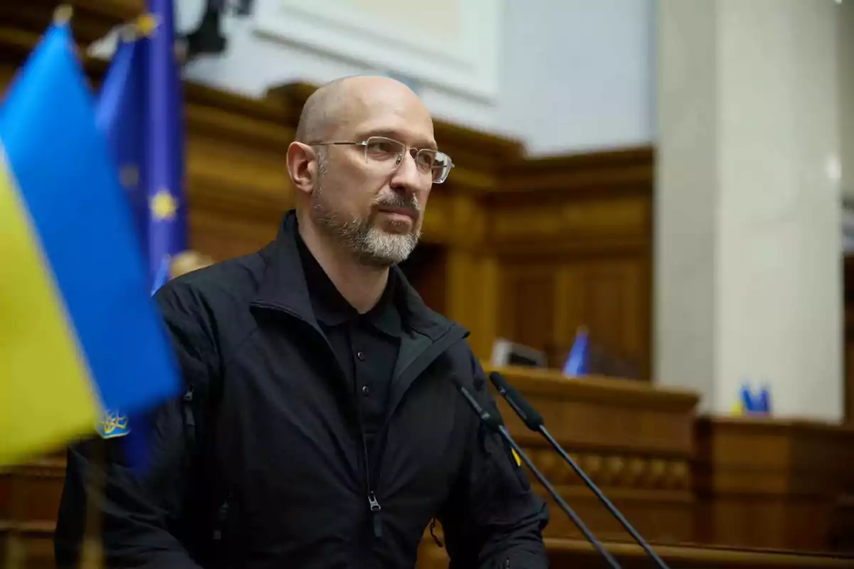 Hombre calvo con barba y gafas hablando en un podio dentro de un edificio gubernamental con banderas de Ucrania y la Unión Europea al fondo Hombre calvo con barba y gafas hablando en un podio dentro de un edificio gubernamental con banderas de Ucrania y la Unión Europea al fondo