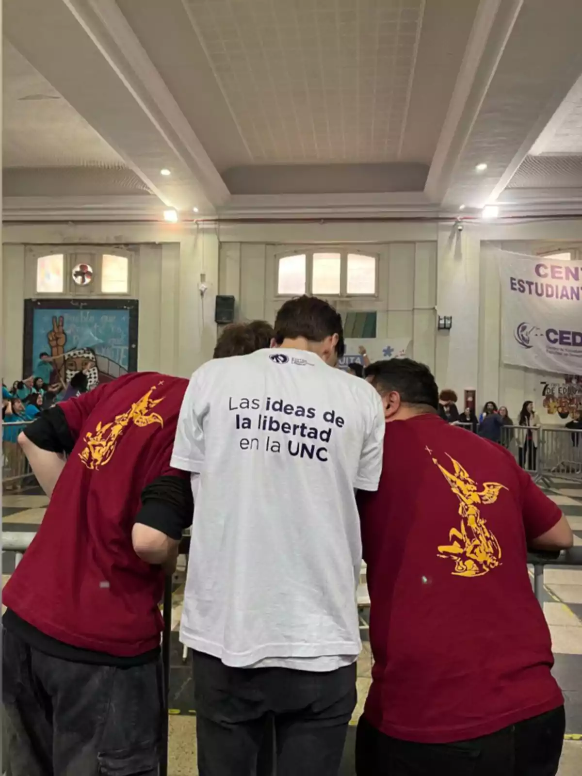 Tres personas de espaldas, dos con camisetas rojas con un diseño amarillo y una con camiseta blanca que tiene un texto en la espalda, dentro de un salón amplio con gente al fondo.