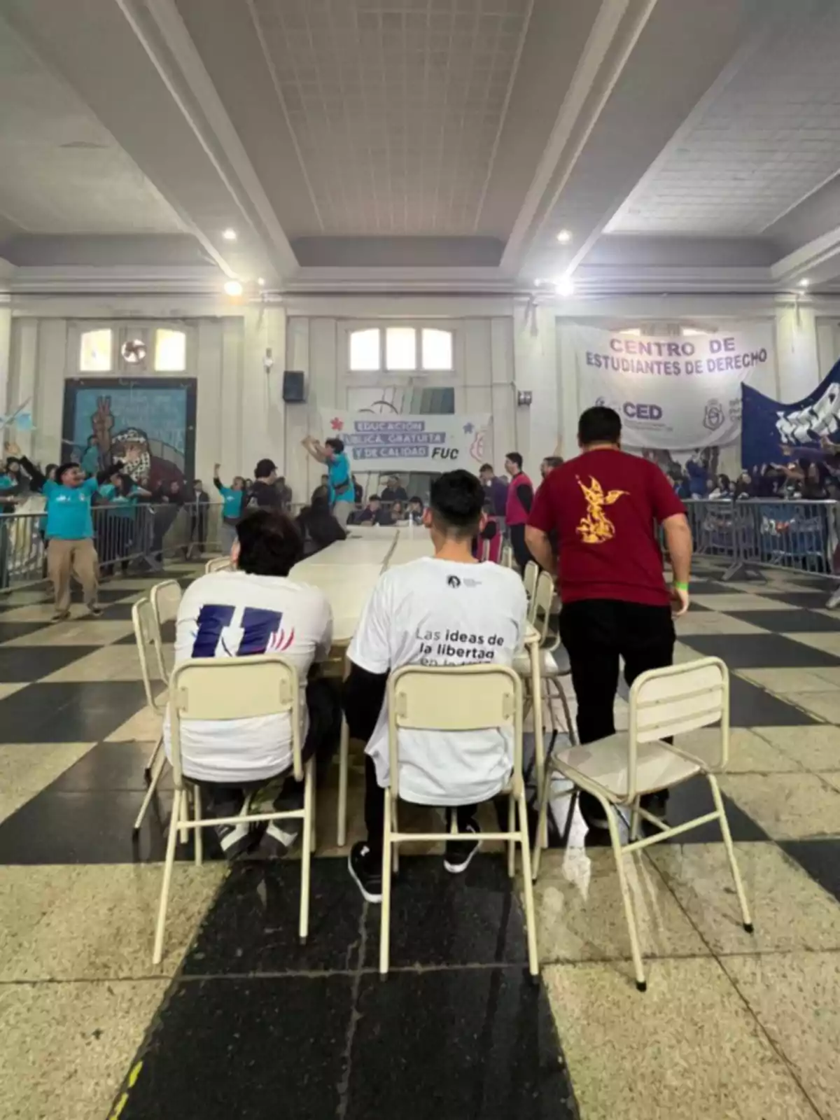 Tres personas sentadas en sillas frente a una mesa en un salón grande con pancartas y público al fondo durante un evento estudiantil