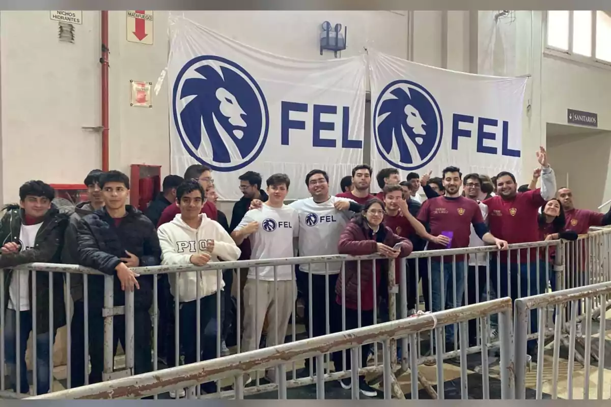 Grupo de jóvenes reunidos detrás de una valla metálica en un evento deportivo o estudiantil con pancartas blancas que tienen el logo de un león azul y las letras FEL