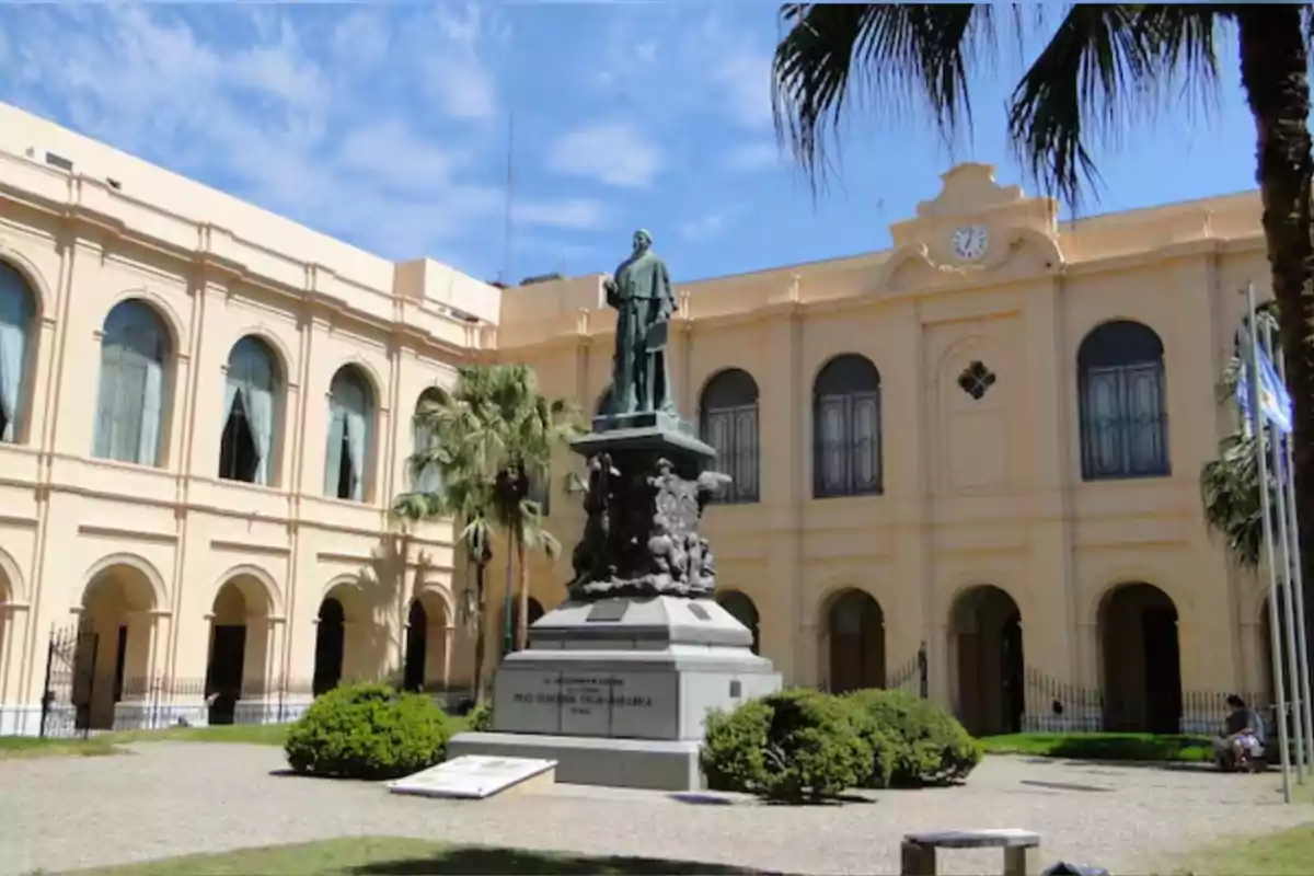 Edificio histórico de color beige con ventanas arqueadas y una estatua en el centro del patio rodeada de palmeras y arbustos bajo un cielo azul