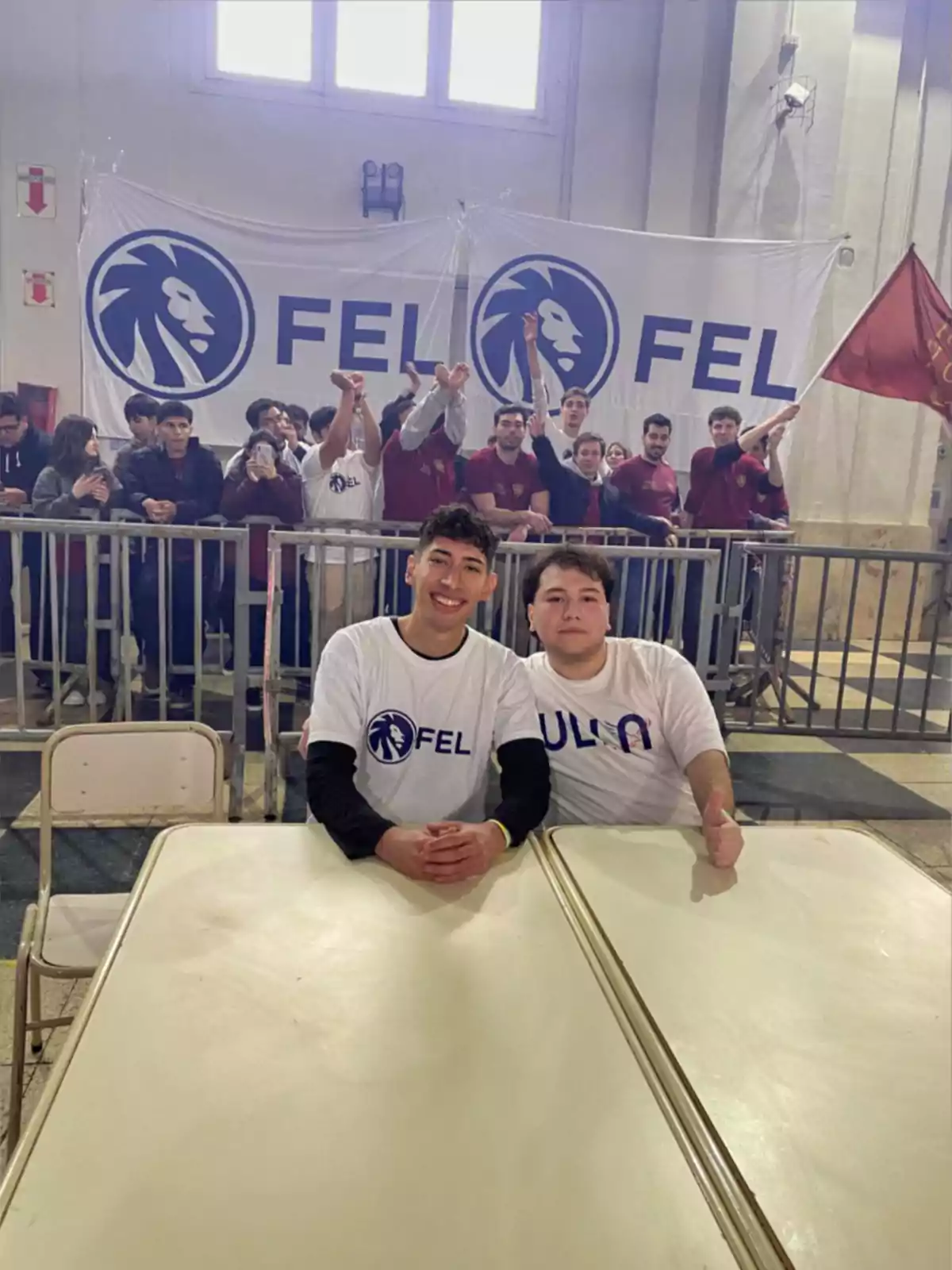 Dos jóvenes con camisetas blancas sentados en una mesa frente a una multitud animada y una gran pancarta con el logo de un león y las letras FEL