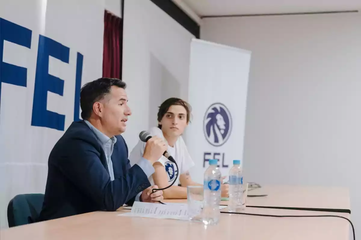 Dos personas sentadas en una mesa durante una conferencia, una de ellas hablando por micrófono y la otra escuchando, con botellas de agua y logotipos de un león en el fondo
