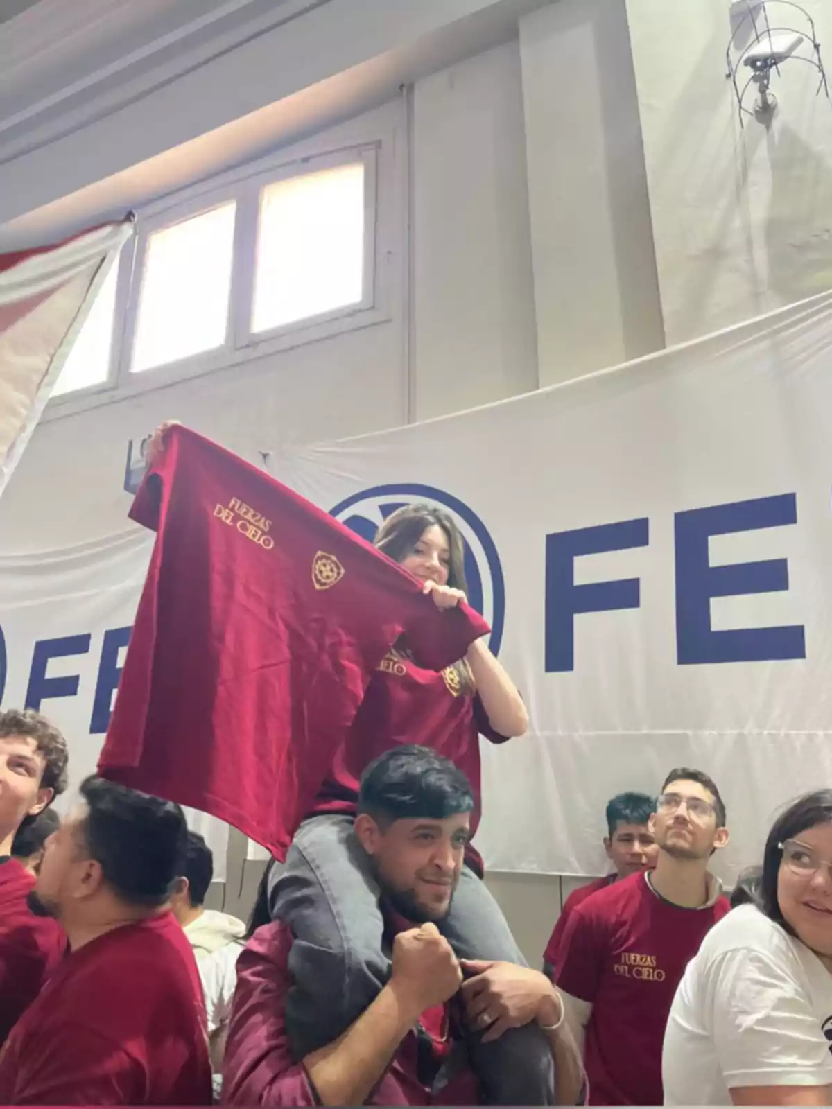 Grupo de jóvenes en un evento, una chica está sentada sobre los hombros de un chico y sostiene una camiseta roja que dice Fuerzas del Cielo, todos lucen animados y llevan camisetas similares