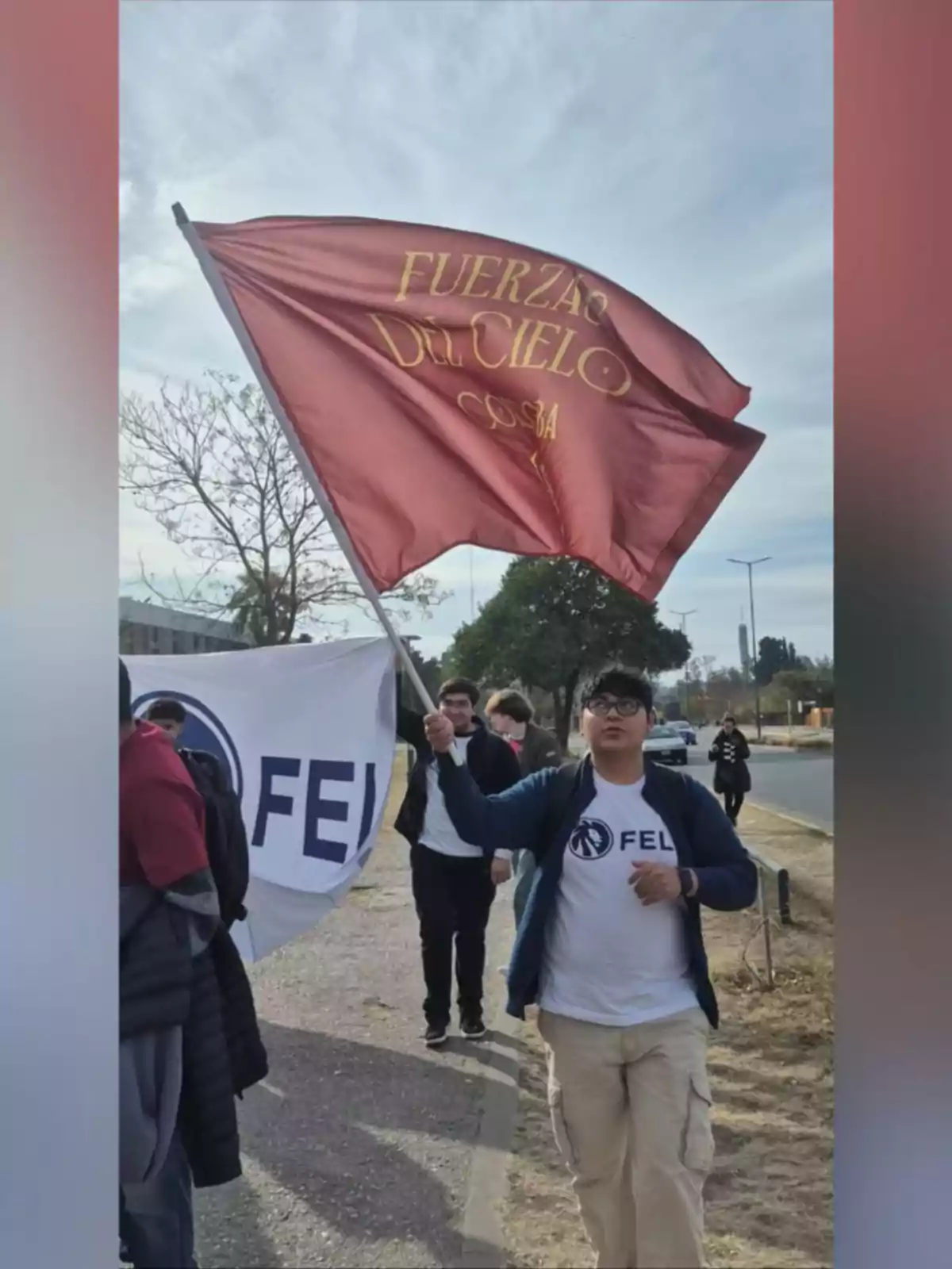 Joven marchando por la calle mientras sostiene una bandera roja con letras amarillas que dice Fuerza del Cielo y viste una camiseta blanca con el logo de FEL acompañado de otras personas