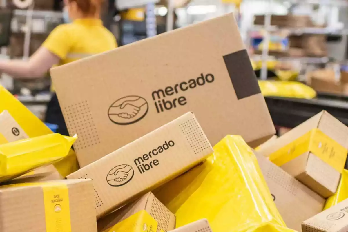 Cajas y paquetes de Mercado Libre apilados en un centro de distribución con una persona desenfocada al fondo